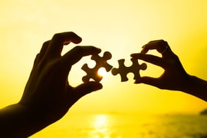 Two hands trying to connect puzzle pieces with sunset background-1
