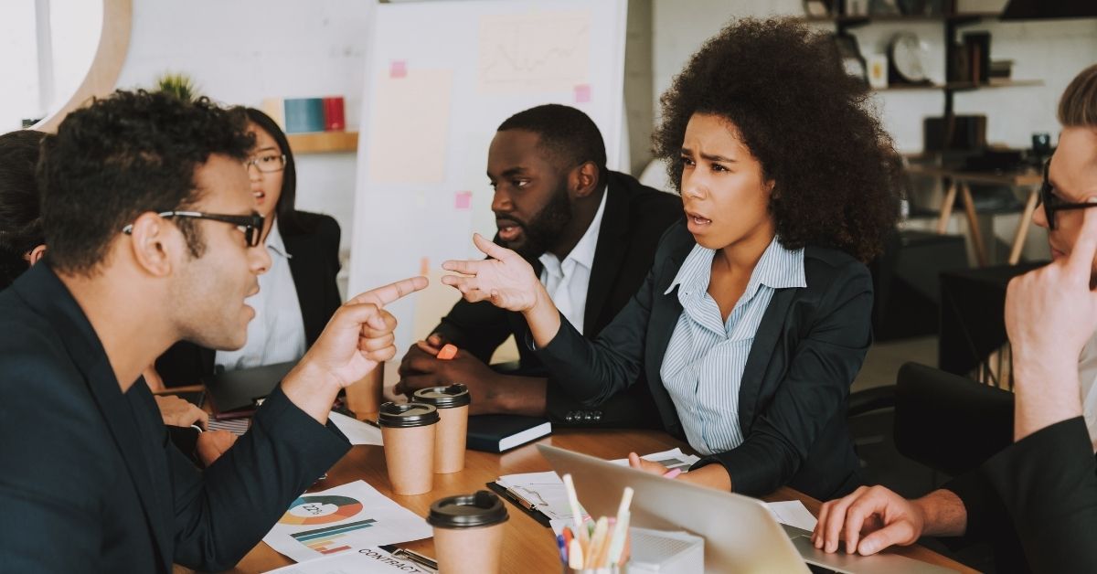 Diverse group of people sitting around a work table, woman upset with a man pointing a finger at her