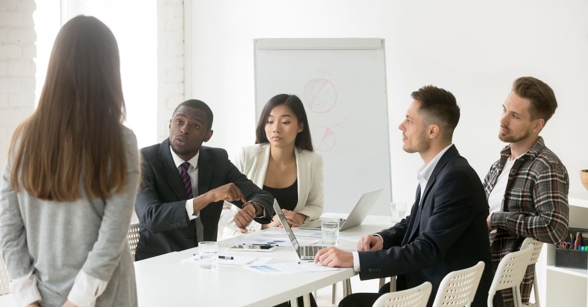 diverse group around a table. many looking at watch scolding someone standing in front of the table