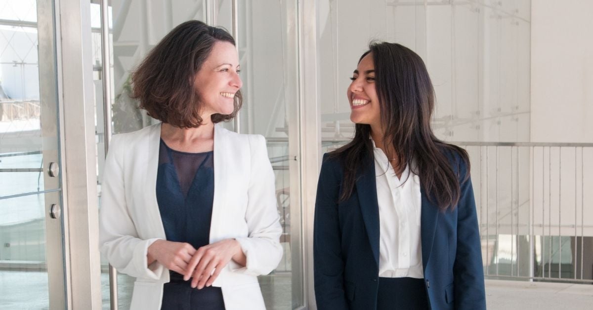 Two professional women walking down a hallway smiling at each other