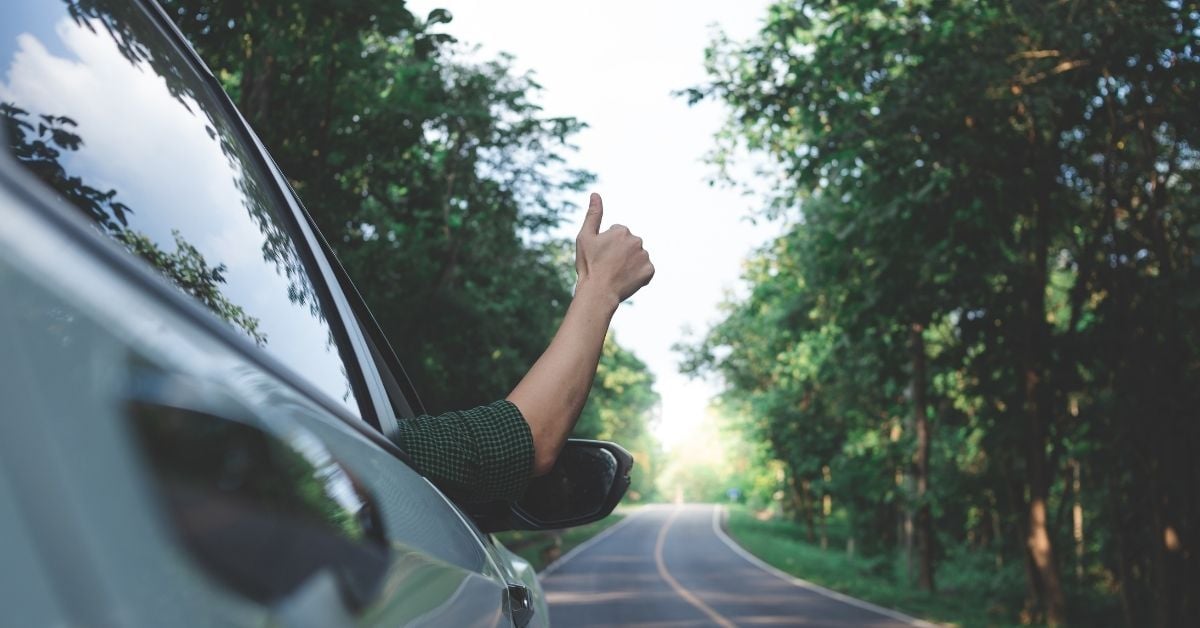 person with their hand out the window in a thumbs up as a car moves down a tree lined highway