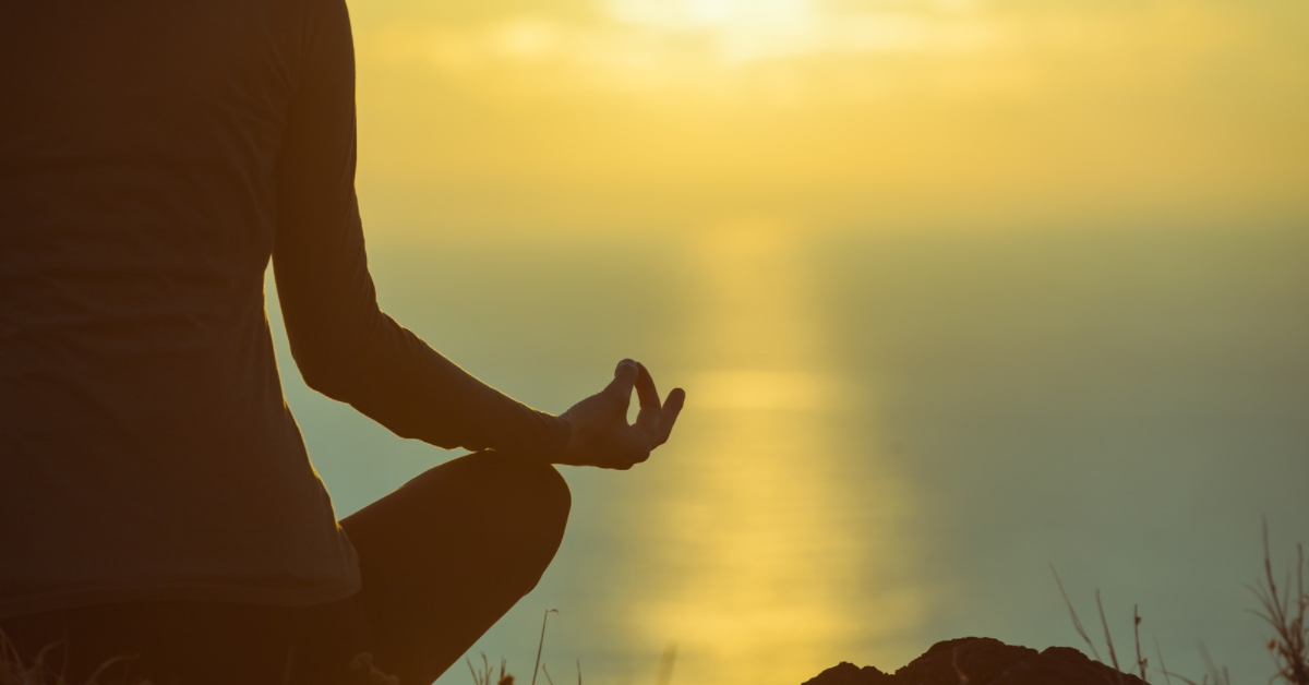 Person sitting meditating in front of a peaceful sunrise, lots of yellows and blues