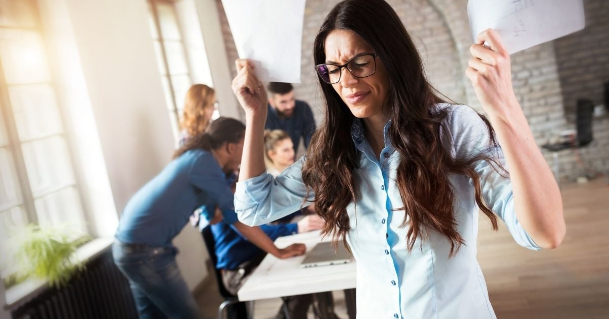 woman holding papers in the air frustrated or overwhelmed with group of coworkers in the background working